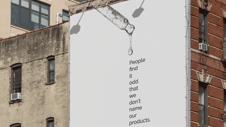 The image shows a large billboard on the side of a building. The billboard is white and features a black and white illustration of a dropper with a drop of liquid hanging from it. The text on the billboard reads "People find it odd that we don't name our products." The billboard is attached to a building with a brick f